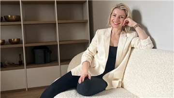 Eine Frau mit blonden Haaren, die einen weißen Blazer und eine dunkle Hose trägt, sitzt auf einer weißen Couch.
