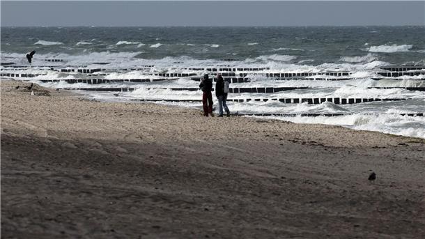 Spaziergänger sind am Ostseestrand bei Graal-Müritz unterwegs, wobei nur ab und zu etwas Sonne durch die grauen Wolken am Himmel fällt.
