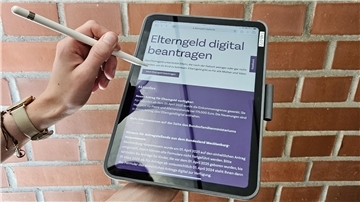 ALT-Text:
Tablet mit geöffneter Webseite „Elterngeld digital beantragen“ vor einer roten Ziegelwand, gehalten von einer Person mit Smartwatch und Stift.
