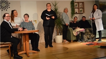 Eine Theatergruppe steht auf einer Bühne in der Kulisse eines Wohnzimmers. Einige Darsteller sitzen an einem Tisch, andere auf einem Sofa, während eine Person in einem weißen Arztkittel steht. 