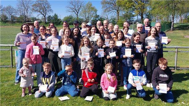 Gruppenfoto vor einem Sportplatz: etwa 30 Teilnehmende aller Altersstufen von Kleinkindern bis Senioren stehen bei sonnigem Wetter auf einer Wiese und halten stolz ihre Urkunden für das Deutsche Sportabzeichen in den Händen.