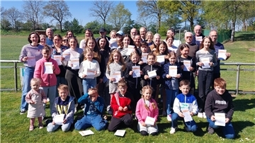Gruppenfoto vor einem Sportplatz: etwa 30 Teilnehmende aller Altersstufen von Kleinkindern bis Senioren stehen bei sonnigem Wetter auf einer Wiese und halten stolz ihre Urkunden für das Deutsche Sportabzeichen in den Händen.
