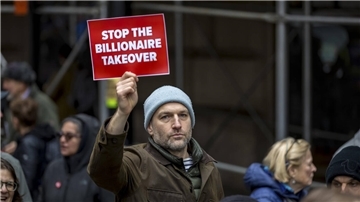 Ein Mann hält ein rotes Schild mit weißer Aufschrift hoch. Darauf heißt es: "Stop the billionaire takeover".  