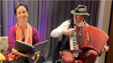 Eine Frau sitzt auf einem Stuhl und hält eine aufgeschlagene Mappe. Neben ihr sitz ein Mann mit einem Akkordeon.