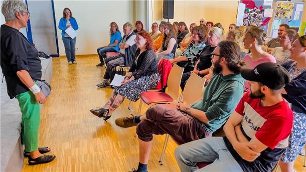 Eine Gruppe von Erwachsenen sitzt in einem Veranstaltungsraum und hört einer Frau im Vordergrund aufmerksam zu. Die Frau steht seitlich zur Kamera und spricht zu den Teilnehmenden. Im Hintergrund sind weitere Personen sowie Plakate und Infowände zu sehen.