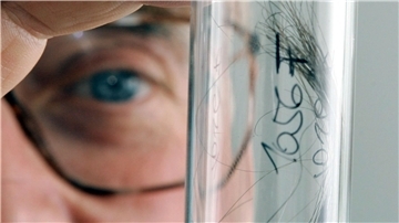 Ein Mann in Nahaufnahme: Man sieht sein Auge hinter einer braunen Brille. Er hält ein Reagenzglas mit einer Haarprobe zwischen den Fingern.