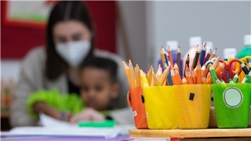 Im Vordergrund bunte Stifte, im Hintergrund eine Frau mit Maske und ein Kindergartenkind.