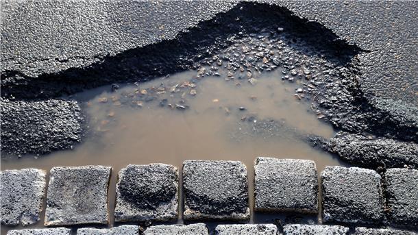 Für Gemeindestraßen: So viel Geld bekommt dein Ort Ein mit Wasser gefülltes Schlagloch in einer Asphaltstraße neben gepflasterten Steinen.