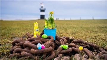 Im Vordergrund liegt eine verrostete schwere Kette, darauf liegt Osterdeko. Im Hintergrund der Leuchtturm "Kleiner Preuße" in Wremen.