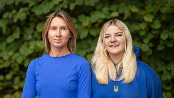 Eine braunhaarige und eine blonde Frau in blauen Oberteilen vor einem Gebüsch.