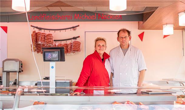 Eine Frau mit roter Jacke. Neben ihr steht ein Mann mit Brille und weißem Kittel. Sie stehen hinter einer Fleischauslage und vor dem Schriftzug "Landfleischerei Michael Neckien".