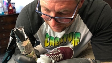 Ein Mann mit einer Brille auf der Nase tätowiert ein Bein.