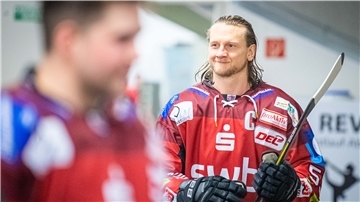 Langhaariger Eishockeyspieler mit rotem Trikot am Körper und Schläger in der Hand.