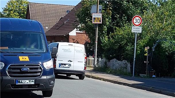 Zwei Lieferwagen fahren auf einer Straße mit Tempo-30-Zone in einem Wohngebiet. Rechts steht ein Verkehrsschild mit der Aufschrift „30 – Straßenschäden“ sowie eine digitale Geschwindigkeitsanzeige, die „22“ km/h zeigt.