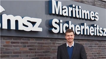 Ein Mann in einem dunklen Anzug und blauen Pullover steht lächelnd vor einer Backsteinwand mit einem Schild, auf dem „Maritimes Sicherheitszentrum“ steht.