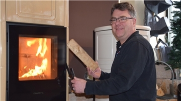 Tino Öltjen sitzt vor einem Kamin. In der rechten Hand hält er ein Stück Holz. 