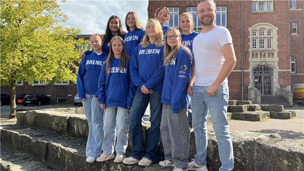 Eine Gruppe Schülerinnen posiert auf einem Schulhof mit dem Lehrer für ein Gruppenfoto.