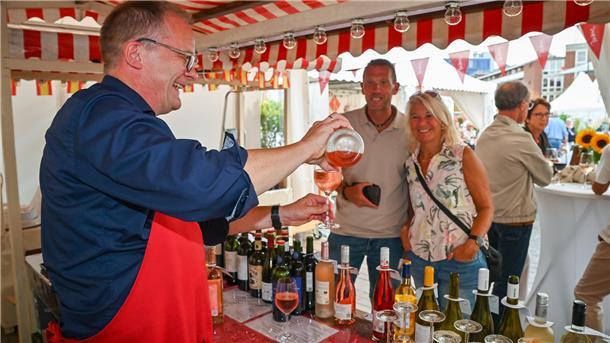 Ein Mann schenkt in einem Verkaufsstand Wein in ein Glas. Im Hintergrund stehen ein weiterer Mann und eine Frau und warten darauf, den Wein in Empfang zu nehmen.