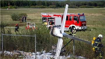 Trümmerteile eines Flugzeugs hängen in einem Zaun, während Einsatzkräfte der Feuerwehr an der Absturzstelle stehen.