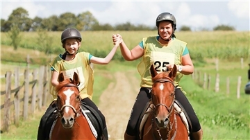 Ein Mädchen und eine Frau sitzen jeweils auf einem braunen Pferd. Sie reiten nebeneinander und halten sich triumphierend die Hände.