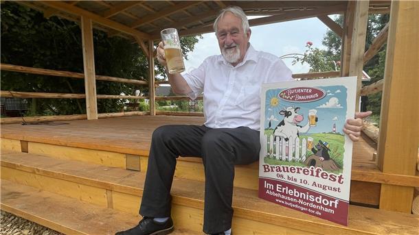 Mann mit Plakat und einem Glas Bier. Gastronom Udo Venema lädt zu einem Brauereifest in sein Erlebnisdorf in Abbehausen ein.