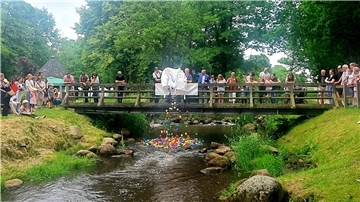 Eine größere Menschengruppe steht auf einer Brücke über einen kleinen Fluss und in den Fluss werden aus einem großen Sack viele bunte Bälle gekippt.