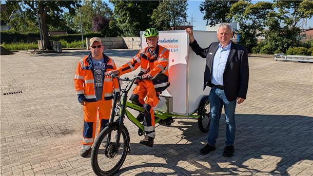 Ein Mann in Arbeitskleidung sitzt auf einem großen E-Lastenrad, zu seiner Rechten steht ein Mann ebenfalls in Arbeitskleidung, zu seiner Linken steht ein Mann in Hemd, Sakko und Jeans. 