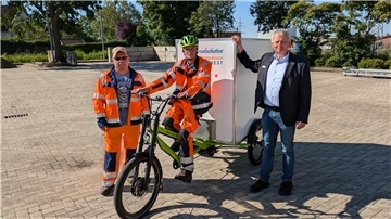 Ein Mann in Arbeitskleidung sitzt auf einem großen E-Lastenrad, zu seiner Rechten steht ein Mann ebenfalls in Arbeitskleidung, zu seiner Linken steht ein Mann in Hemd, Sakko und Jeans. 