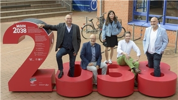 Fünf Personen stehen und sitzen um die rote Installation „MOIN 2038“ auf dem Campus der Hochschule Bremerhaven, die symbolisch für das Klimaziel des Landes Bremen steht.