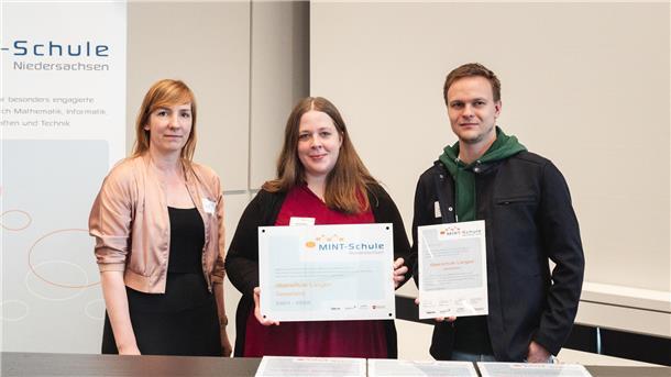 Zwei Frauen und ein Mann stehen nebeneinander. Sie halten ein Schild "MINT-Schule" und eine Urkunde hoch. 