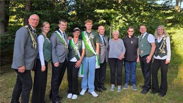 Eine Gruppe von Schützen in festlicher Kleidung und mit Schärpen sowie Orden steht nebeneinander im Freien. Männer und Frauen verschiedener Altersgruppen posieren lächelnd für das Foto vor einer Kulisse aus Bäumen und Grün.