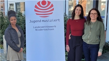 Zwei junge Frauen und eine Erwachsene stehen vor einem Gebäude neben einem Banner mit der Aufschrift „Jugend musiziert – Landeswettbewerb Niedersachsen“.