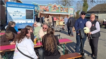 Ein fröhliches Marktgeschehen bei sonnigem Wetter: Menschen sitzen an Biertischen, genießen Speisen und Getränke. Im Hintergrund ein blauer Verkaufswagen „Krabbenkutter Polli“ sowie ein bunt dekorierter Süßwarenstand mit Lebkuchenherzen. Zwei Personen stehen lächelnd mit Luftballons neben einem E-Scooter.