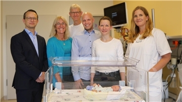 Sechs Erwachsene, darunter medizinisches Personal und weitere Personen, stehen lächelnd in einem Krankenhauszimmer hinter einem Inkubator mit der Puppe eines neugeborenen Babys darin.