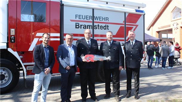 Fünf Männer, darunter Feuerwehrleute in Uniform und Vertreter der Gemeinde, stehen vor einem neuen Feuerwehrfahrzeug mit der Aufschrift „Feuerwehr Bramstedt“. Einer der Feuerwehrmänner hält symbolisch einen großen, verpackten Fahrzeugschlüssel. Im Hintergrund sind Besucherinnen und Besucher beim Tag der offenen Tür an der Fahrzeughalle zu sehen.