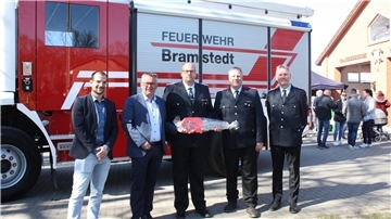 Fünf Männer, darunter Feuerwehrleute in Uniform und Vertreter der Gemeinde, stehen vor einem neuen Feuerwehrfahrzeug mit der Aufschrift „Feuerwehr Bramstedt“. Einer der Feuerwehrmänner hält symbolisch einen großen, verpackten Fahrzeugschlüssel. Im Hintergrund sind Besucherinnen und Besucher beim Tag der offenen Tür an der Fahrzeughalle zu sehen.
