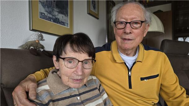 Eine ältere Frau und ein älterer Mann schauen in die Kamera, der Mann hat den Arm um die Schulter der Frau gelegt.