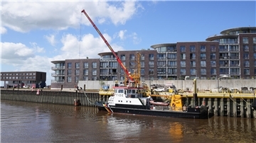Eine Uferpromenade: An einer Kaje wird mithilfe eines Krans gearbeitet. Daneben: Ein Schiff.