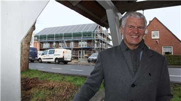 Ein Mann steht unter einem Pavillon auf einer Grünfläche und lächelt in die Kamera. Im Hintergrund steht ein eingerüstetes Haus.