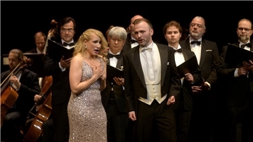 Zwei Solisten singen im festlichen Bühnenoutfit vor einem Chor und Orchester in schwarzer Abendgarderobe während einer Operettengala.