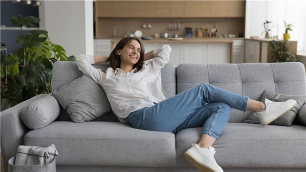 Eine dunkelhaarige Frau mit weißer Bluse und Jeans sitzt auf einem grauen Sofa und hat die Arme hinter dem Kopf verschränkt.