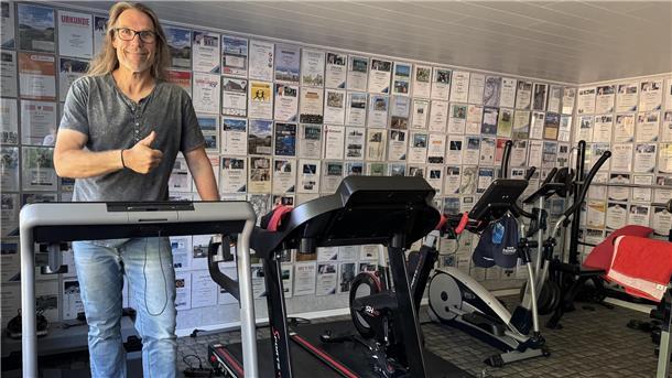 Ein langhaariger Mann mit Brille und Jeans in einem T-Shirt steht in einem privaten Fitnessstudio auf einem Laufband.