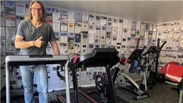 Ein langhaariger Mann mit Brille und Jeans in einem T-Shirt steht in einem privaten Fitnessstudio auf einem Laufband.