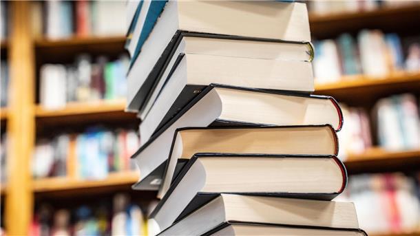 Ein hoher Stapel Bücher mit festen Einbänden steht im Vordergrund, während im Hintergrund unscharf Bücherregale mit weiteren Büchern zu sehen sind. Die Bücher sind unterschiedlich groß und haben verschiedenfarbige Einbände.