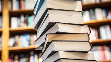 Ein hoher Stapel Bücher mit festen Einbänden steht im Vordergrund, während im Hintergrund unscharf Bücherregale mit weiteren Büchern zu sehen sind. Die Bücher sind unterschiedlich groß und haben verschiedenfarbige Einbände.