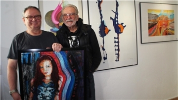 Zwei Männer stehen in einer Galerie nebeneinander und halten ein gerahmtes Gemälde mit einer farbintensiven Frauenfigur. Hinter ihnen hängen mehrere abstrakte Bilder an der weißen Wand.