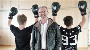 Ein Mann steht zwischen zwei Jugendlichen, die Boxhandschuhe tragen.