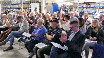 Zahlreiche Erwachsene sitzen dicht an dicht auf Stuhlreihen in einer Werkhalle und applaudieren. Im Hintergrund sind Regale mit Kisten sowie Werkbänke zu sehen.