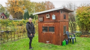 Frau im Garten neben einem Gartenhaus, auf dem "Igelhaus" steht.  