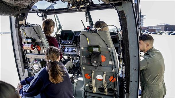 Mehrere Mädchen sitzen im Cockpit eines Militärhubschraubers und schauen interessiert auf die Instrumente; ein Soldat in Fliegerkombi erklärt technische Details und steht seitlich an der offenen Tür.